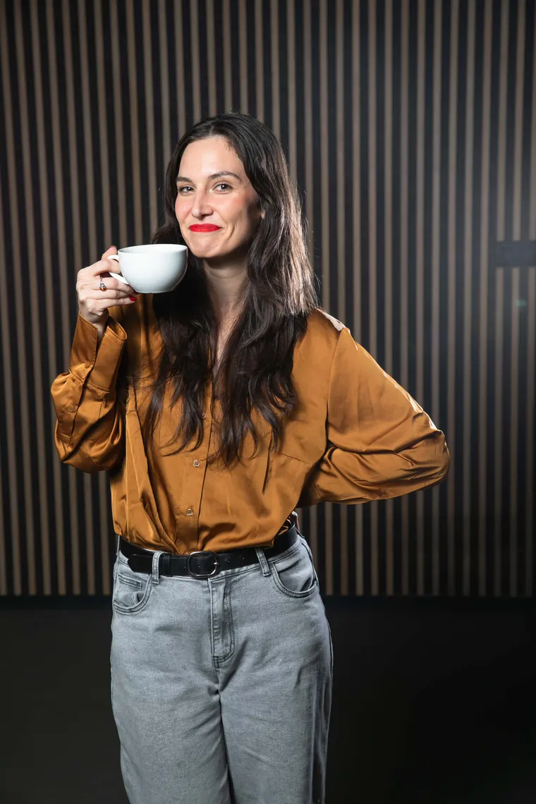 Tess in an ochre shirt holding a cup of coffee, ready for a chat